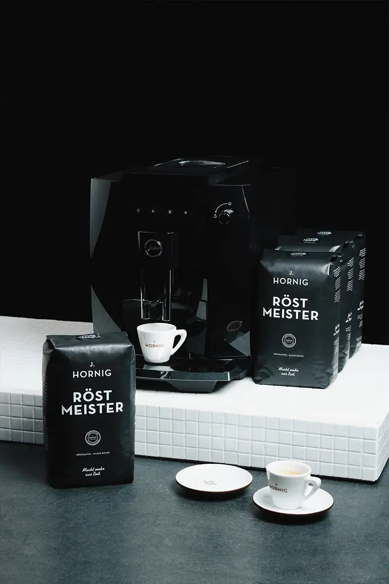 Vier schwarze Packungen J. Hornig Röstmeister Kaffee und ein Kaffeevollautomat auf weißem Untergrund.