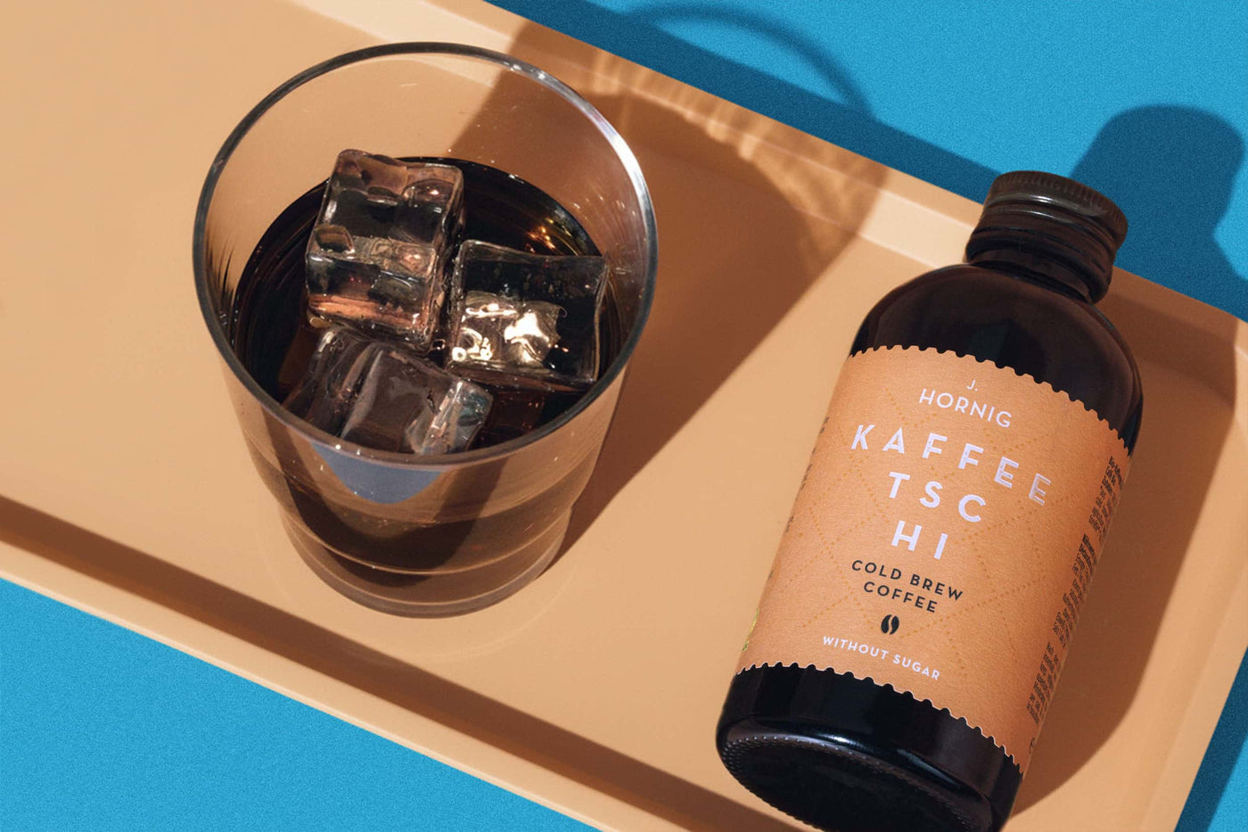 J. Hornig Kaffeetschi Cold Brew Flasche neben Glas mit Eiswürfeln auf beigem Tablett, blauer Hintergrund.