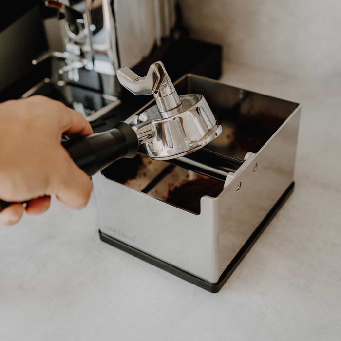 Hand hält einen Siebträger über einer Edelstahl-Knockbox, um Kaffeepuck auszuklopfen.
