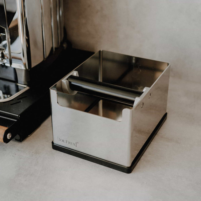 Edelstahl-Knockbox von JoeFrex mit Gummibalken auf heller Küchenarbeitsfläche neben einer Espressomaschine.