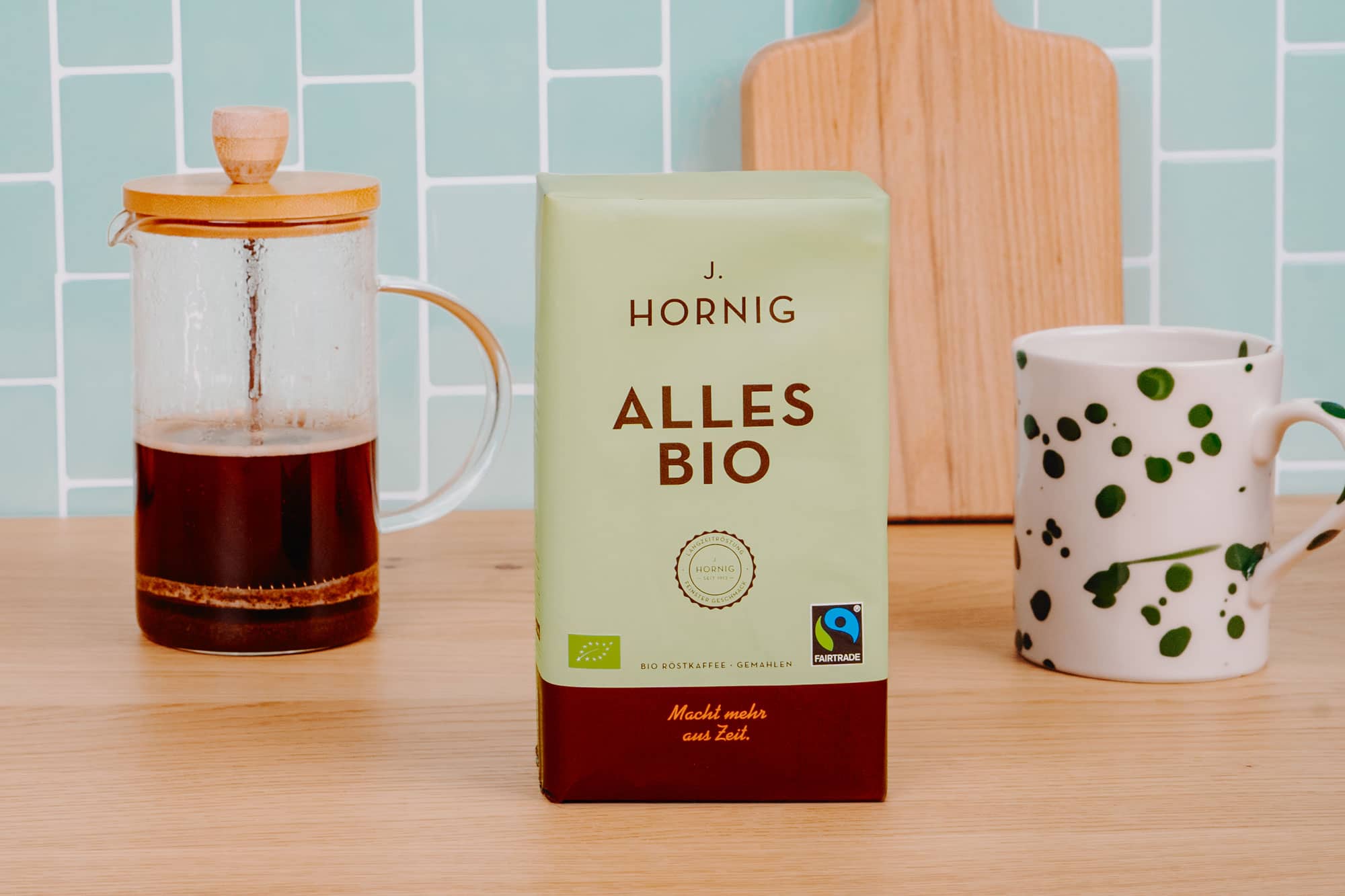 J. Hornig Alles Bio Kaffeepackung auf Holzarbeitsfläche, links eine Glaskaraffe mit Kaffee, rechts eine gemusterte Tasse, im Hintergrund grüne Fliesenwand und Holzbrett.