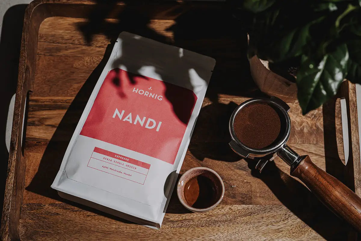Eine Packung J. Hornig Nandi Spezialitätenkaffee Espresso auf einem Holztablett mit einem Siebträger und einer Espressotasse.