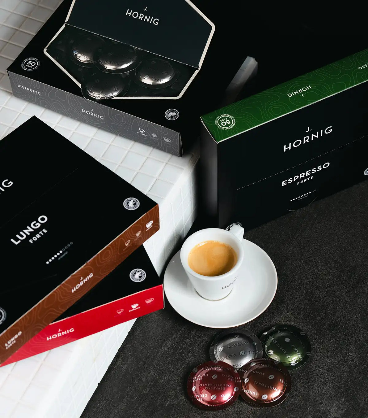 Vier Packungen J. Hornig Pro Pads Kaffee mit einer Kaffeetasse auf schwarzem und weißem Untergrund.