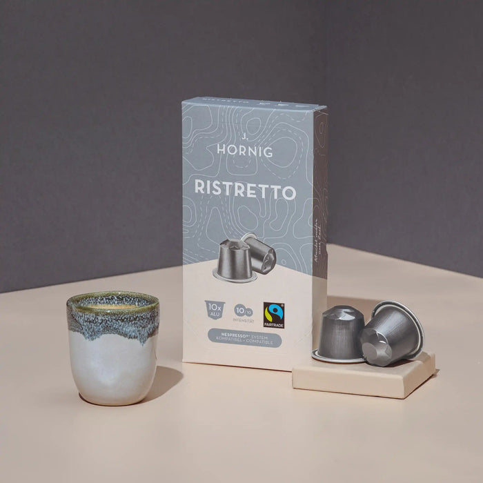 Eine Packung J. Hornig Ristretto Kaffeekapseln mit zwei Alukapseln und einer Kaffeetasse auf hellem Untergrund mit einem grauen Hintergrund
