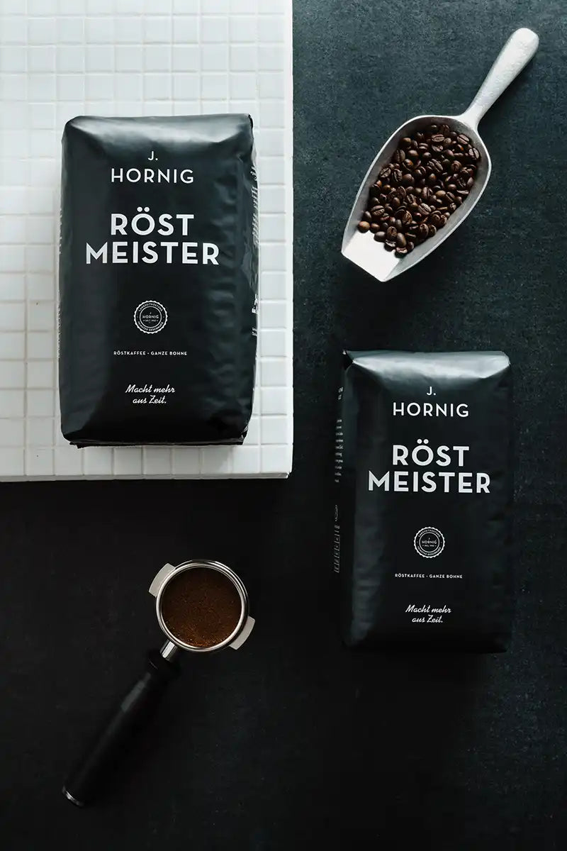 Zwei Packungen J. Hornig Röstmeister mit einer Schaufel voller Kaffeebohnen und einem Siebträger auf schwarzen Hintergrund.