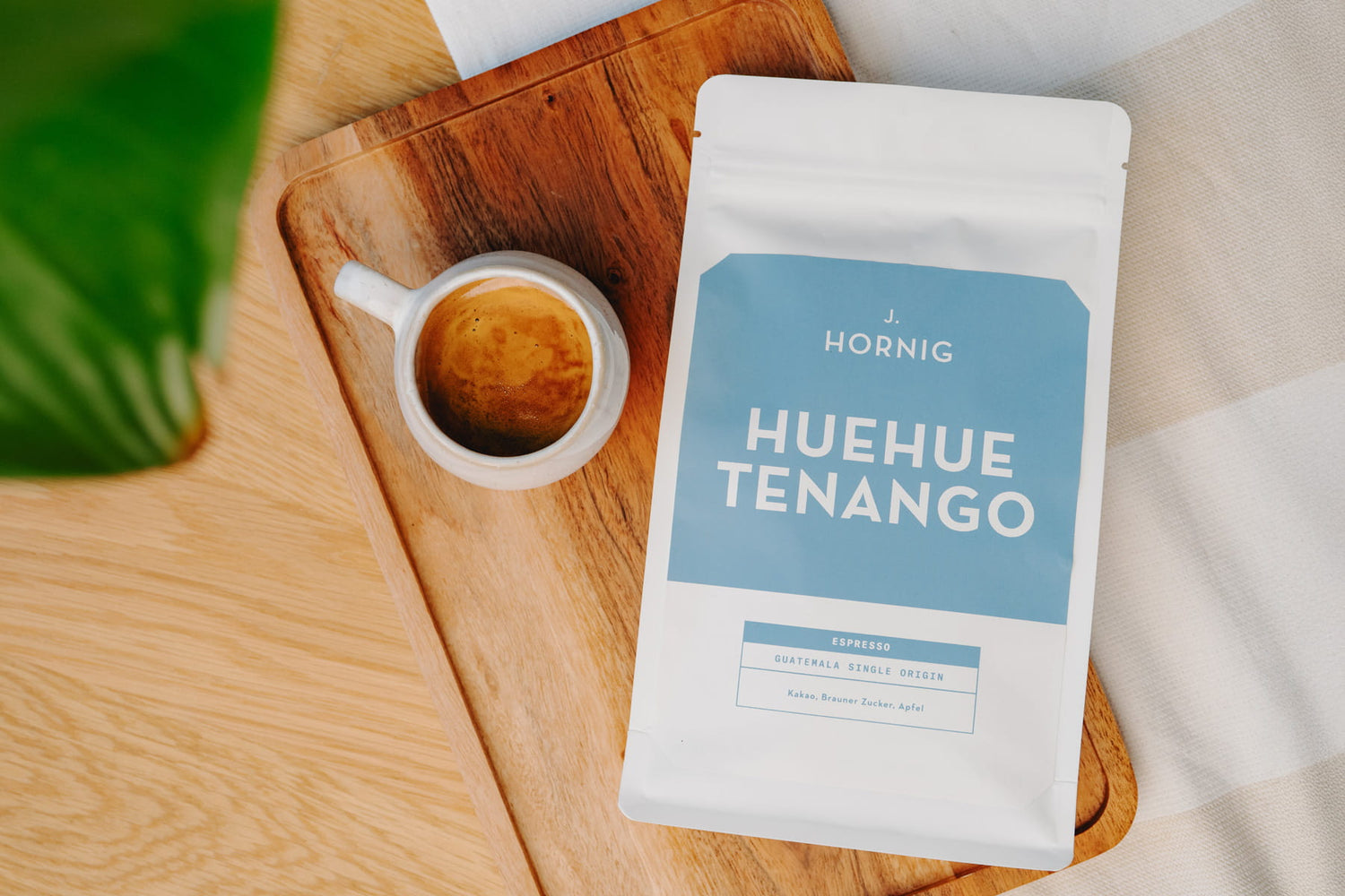 Packung J. Hornig Huehuetenango Spezialitätenkaffee neben einer Tasse Kaffee auf einem Holzbrett mit grünem Blatt im Hintergrund.