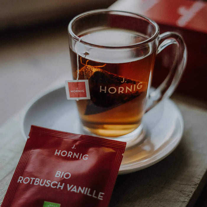 Eine Tasse Tee in einem J.Hornig Teeglas mit einer Einzelpackung Bio Vanille Rotbusch Tee.