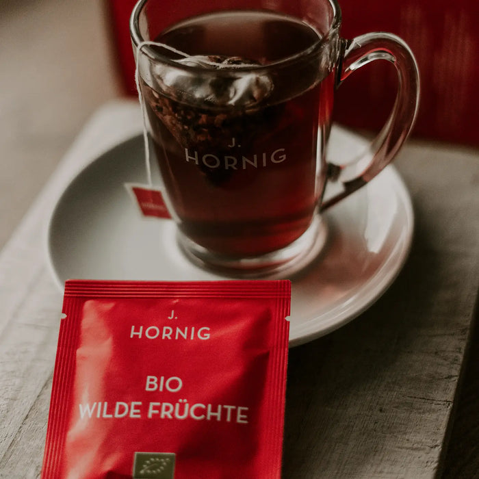 Eine Tasse Tee in einem J.Hornig Teeglas mit einer Einzelpackung Bio Wilde Früchte Tee.