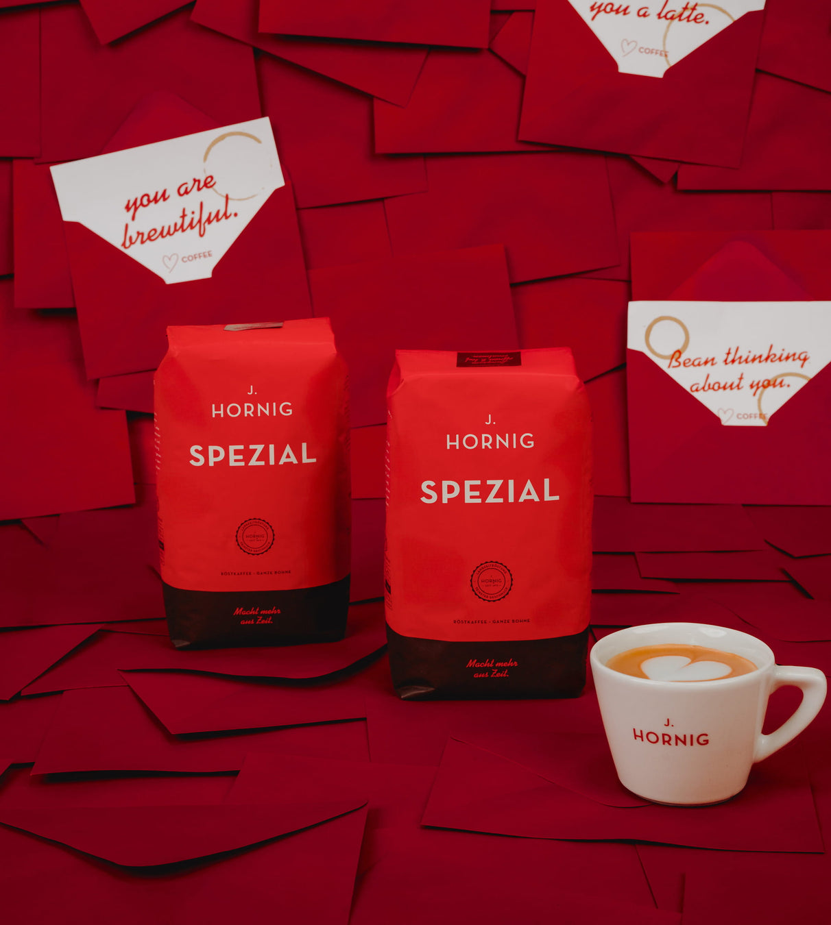 Zwei Packungen J. Hornig Spezial Kaffee auf rotem Papierhintergrund, davor eine weiße J.-Hornig-Espressotasse mit Kaffee, im Hintergrund mehrere rote Kuverts und Grußkarten.