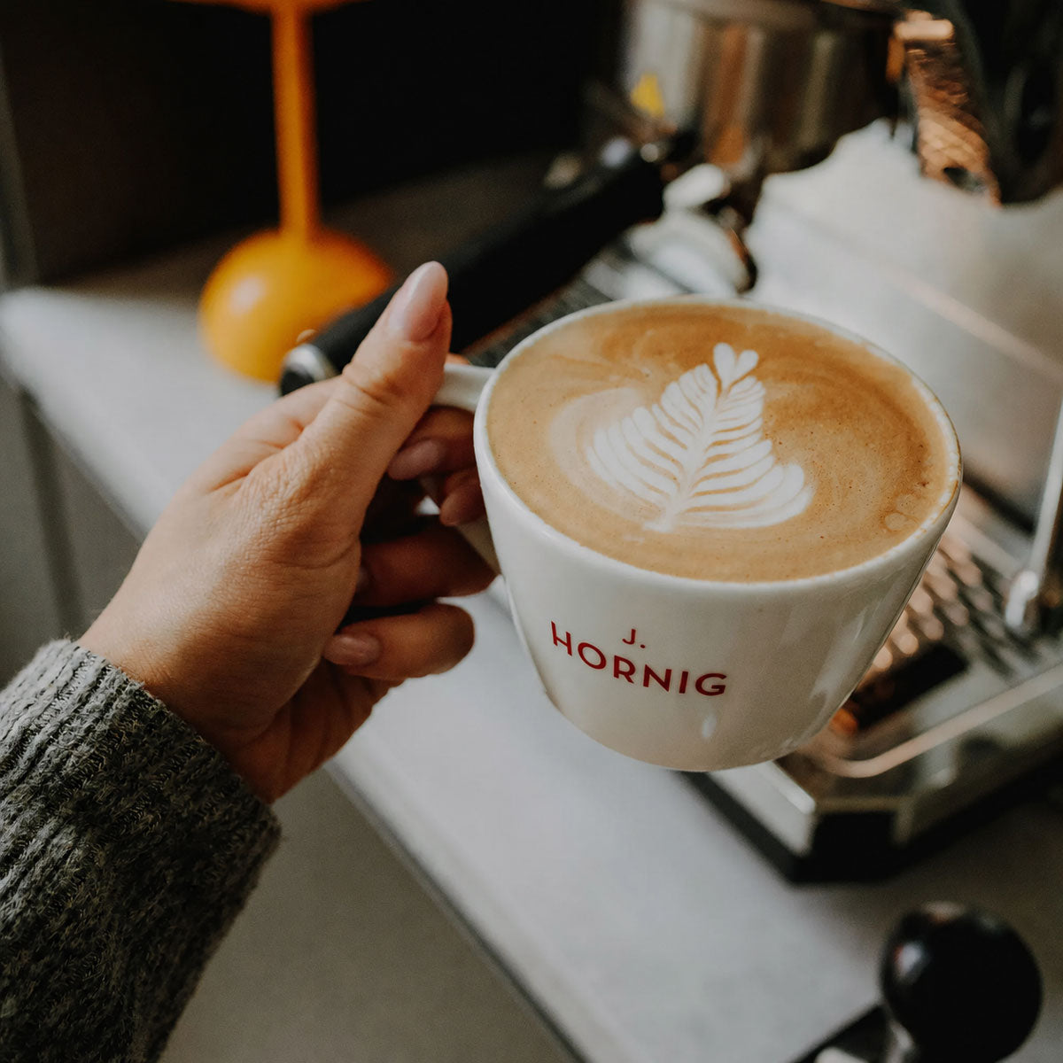 Eine Hand hält eine J. Hornig Tasse Cappuccino mit Latte Art.