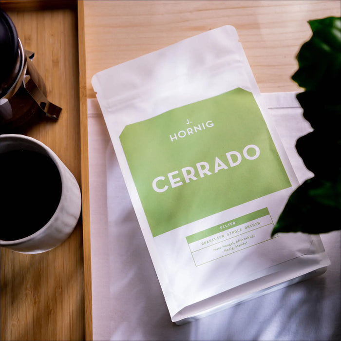 Eine Packung J. Hornig Spezialitätenkaffee Cerrado Filter liegend mit einer gefüllten Kaffeetasse und einer Zimmerpflanze auf einem Holztisch mit einem weißen Tischtuch
