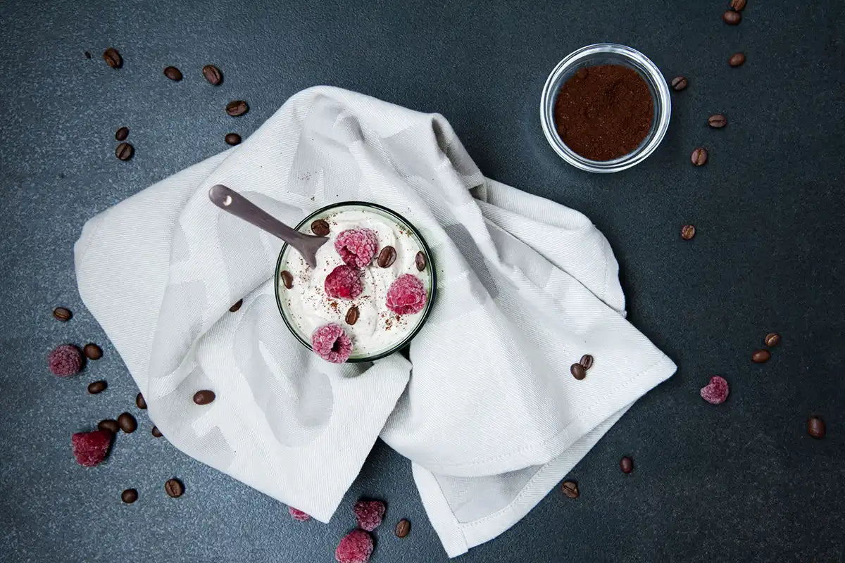 Cremiges Kaffee Joghurt mit gefrorenen Himbeeren und Kaffeebohnen von oben auf einem weißen Tuch 