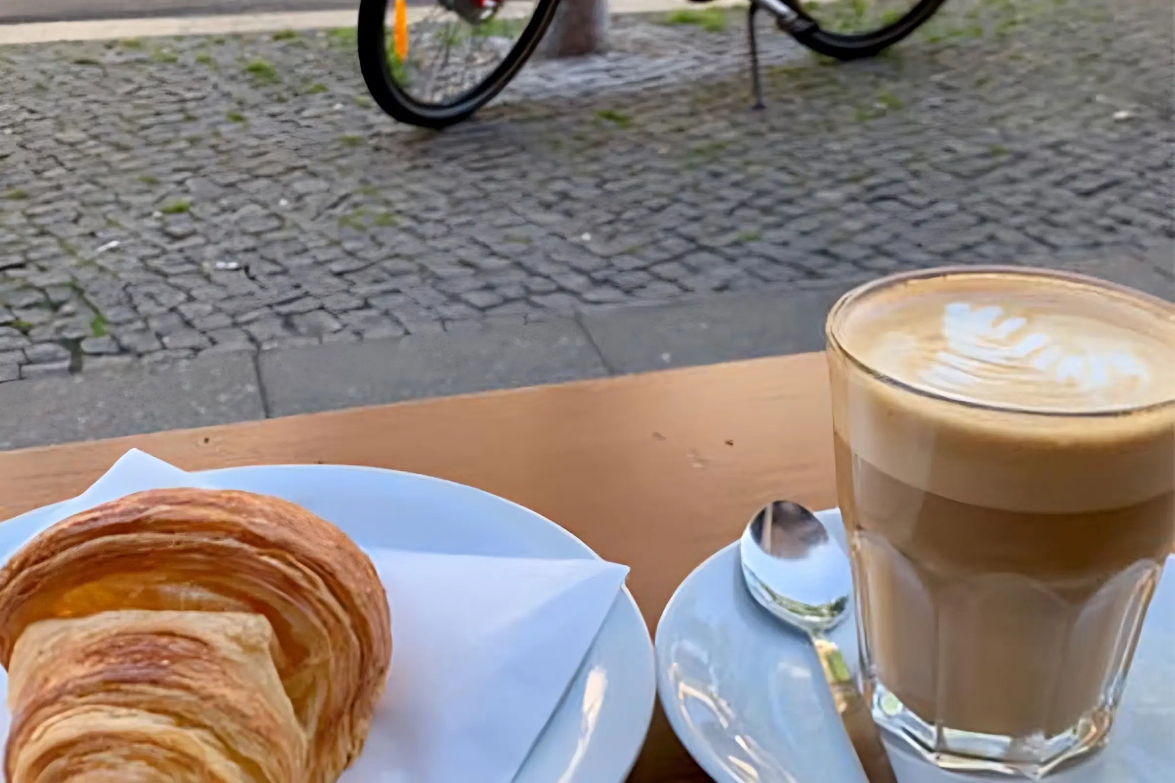 Ein Caffe Latte und Croissant sind im Vordergrund dahinter eine Straße mit einem roten Fahrrad 