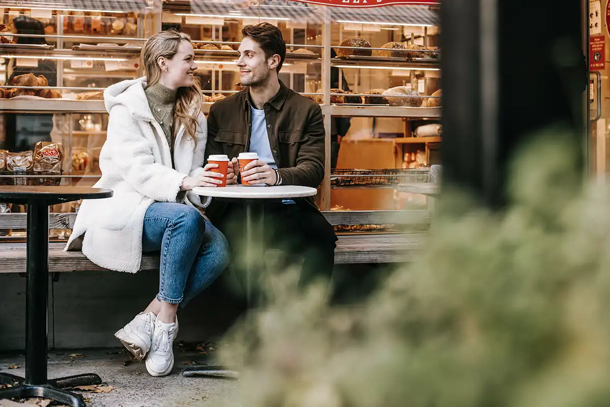 Ein Paar sitzt vor einem Coffeeshop mit roten Coffee-To-Go Bechern und sieht sich an
