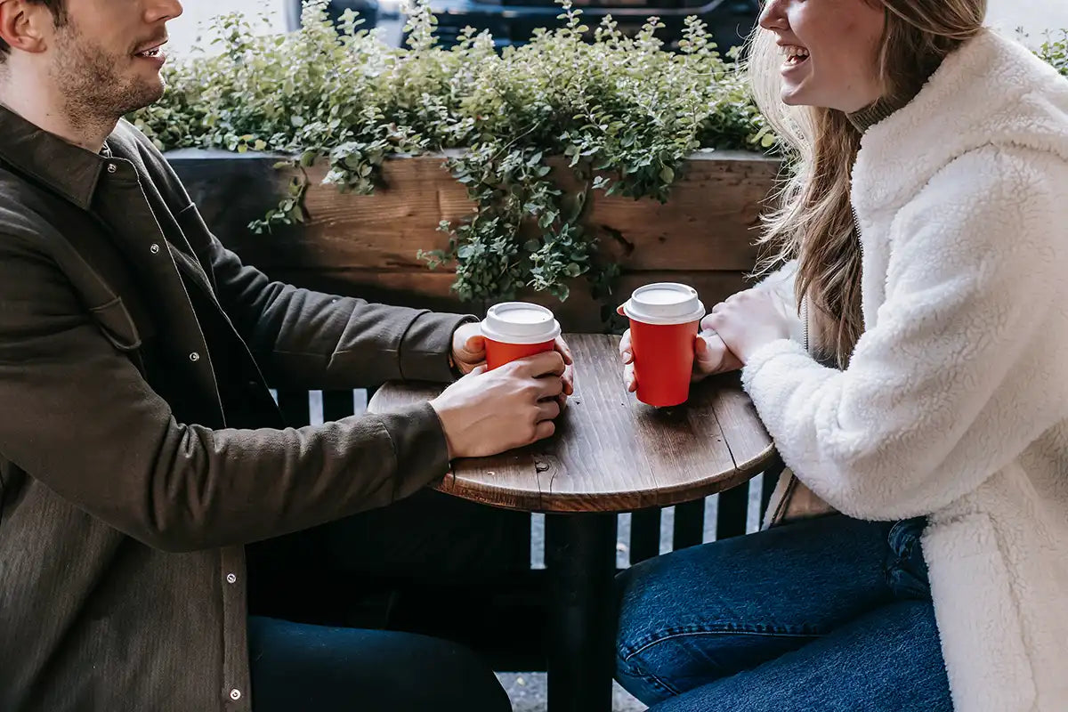 Ein Paar sitzt sich mit roten Coffee-To-Go Bechern gegenüber und lacht sich an 